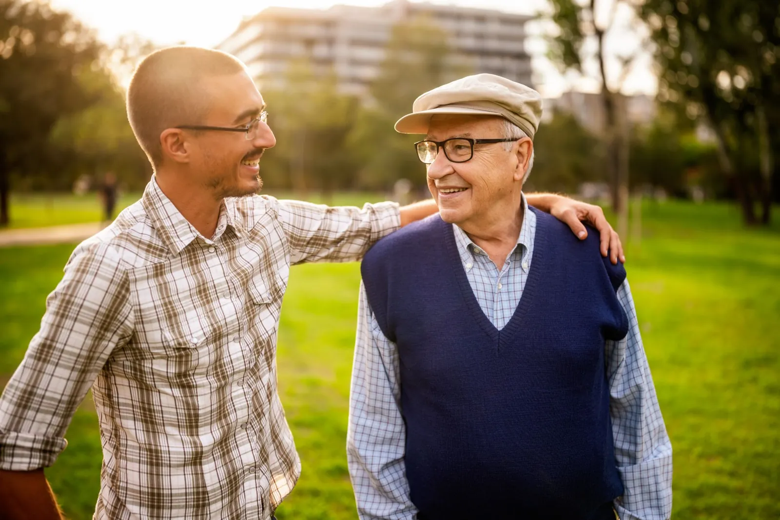 A younger man rests his arm on an older man's shoulder as they share a warm smile in a sunlit park