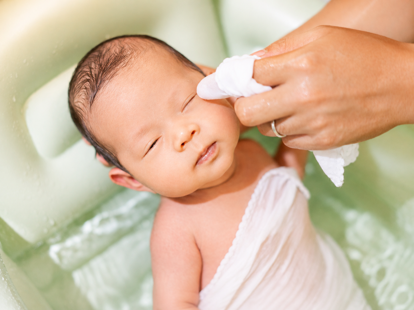 A newborn being gently cleaned with a plain white cloth during a bath, illustrating that most babies need only water and a cloth rather than a routine of scented products